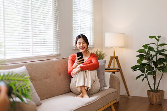 Happy young asian woman relax on comfortable couch at home texting or chatting messaging on the phone, smiling girl use cellphone, online message, shopping online from home
