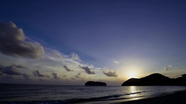 bright yellow sun descends behind pu island as soft blue clouds drift in the sky at kata beach phuket beautifully captured in time lapse to show the evening&rsquo;s color transformation