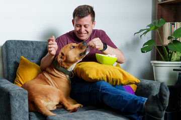 Happy man feeding popcorn to his dog while relaxing and watching television on the sofa in living room