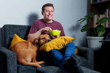 Young man relaxing on sofa with his dog, enjoying popcorn while watching television at home