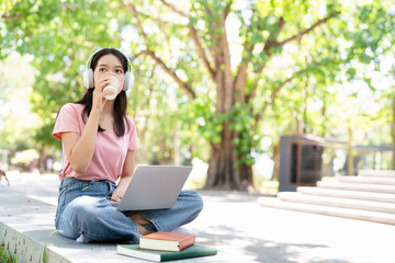 Young female student with laptop and headphones studying online sitting outdoor drinking coffee at...