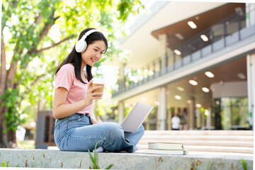 Young female student with laptop and headphones studying online sitting outdoor drinking coffee at...