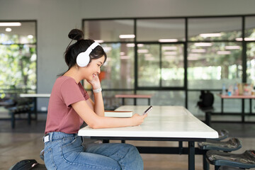 Young student woman wear casual clothes headphones sits alone in cafeteria  listen music use mobile...