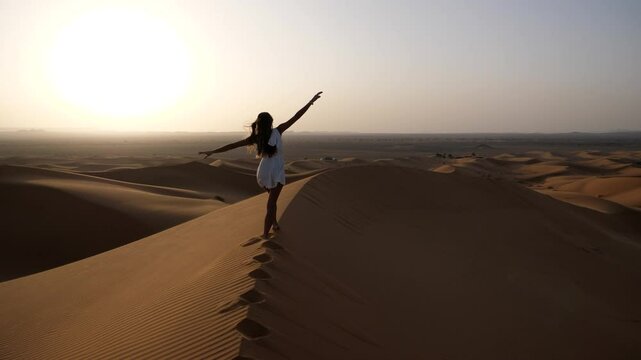 Woman walking barefoot along the sharp crest of a sand dune in Erg Chebbi desert during a stunning sunset in Morocco, surrounded by endless golden dunes and peaceful silence