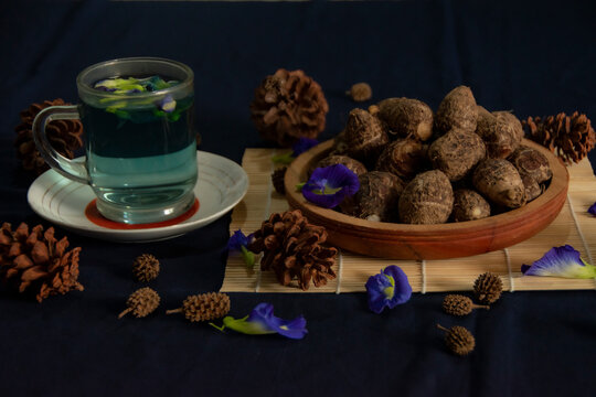 A cup of clitoria ternatea tea served beside a bowl of steamed eddoe roots, styled with pinecones and butterfly pea flowers on a rustic bamboo mat.