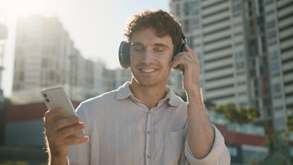 Happy positive Caucasian man holding mobile phone smartphone listening music in headphones guy...
