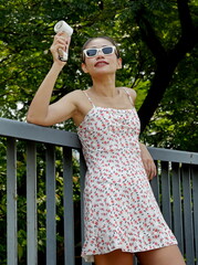 An Asian woman relaxes in the shade on a bridge, holding a portable fan, wearing a floral dress...