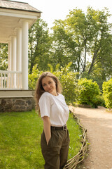 Young woman standing calmly in the garden of an old estate. Casual outfit and relaxed pose contrast beautifully with the historical architecture and lush greenery of a sunny summer day.