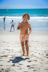 Smiling young African American boy running and playing at the beach while on a family vacation. Playing in the ocean with other people enjoying the beach in the background