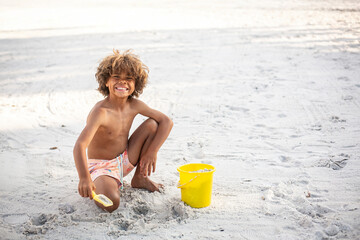 Cute diverse little boy playing in the sand at the beach while on vacation. Digging in the sand with a shovel and bucket