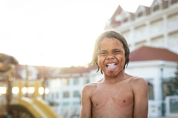 A sassy, diverse young boy playing in a resort pool. Wet and have a great time as he sticks out his tongue and shows a funny expression on his face. Fun waterpark lifestyle photo