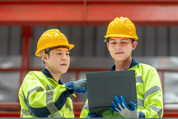 Two construction workers wea yellow safety helmets and high-visibility jackets discussing project details while using a laptop at an industrial site