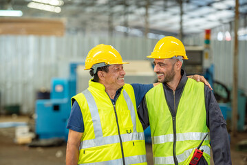 Two happy workers in hard hats and safety vests embrace in a factory.