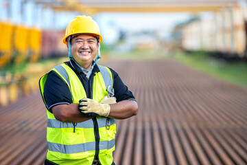 Confident Asian Engineer with Crossed Arms at Railway Construction Site Smiling Foreman in Hard Hat: Professional Worker at Train Depot Proud Industrial Technician Standing on Railway Tracks.