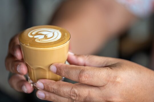 Close-up of hands holding a glass of creamy coffee latte with latte art heart design on foam in a cozy cafe setting, warm beverage mood, detailed shot