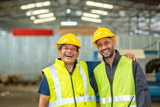 Two happy factory workers wearing hard hats and safety vests standing with arms crossed.