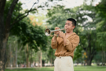 Young man playing trumpet in a park surrounded by lush green trees on a bright day