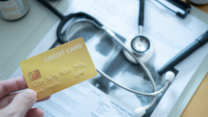 A hand holding a black credit card above stethoscope and medical records on a desk.