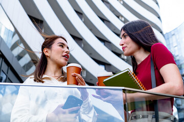 Two businesswomen holding takeaway coffee cups and notebooks are having a conversation during their...