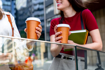 Two businesswomen are holding disposable coffee cups while smiling and chatting during a break...