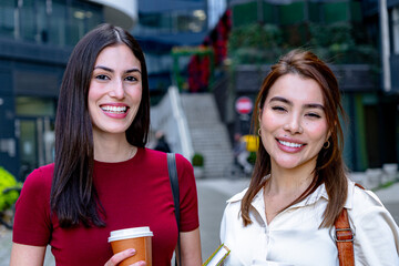Two young businesswomen are smiling and looking happy while holding a coffee and a notebook in their hands, in the city
