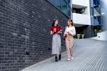 Two female professionals walking outside office building holding takeaway coffee and discussing work