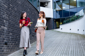 Two young businesswomen holding takeaway coffee and notebooks are walking and chatting outside modern office buildings