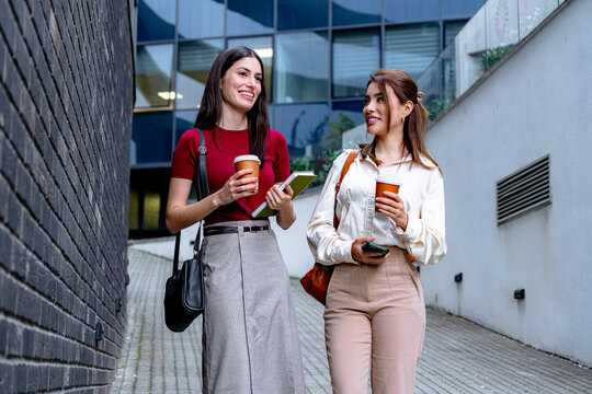 Two businesswomen walking in the city during coffee break, holding takeaway coffees and notebook and smartphone