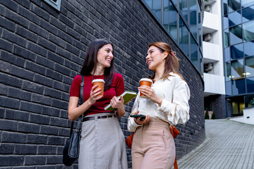 Two young businesswomen smiling and talking during coffee break near office building, holding notebooks and smartphones