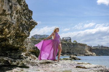 Woman dress sea: Blonde in purple dress poses on rocky coast during daytime, enjoying the scenic...
