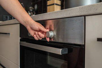 Close-up of a woman's hand adjusting a silver control knob on a modern built-in kitchen oven with a...