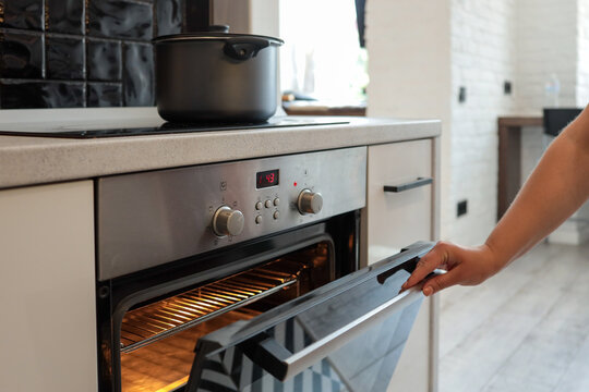 Close-up of a hand opening a hot oven in a modern kitchen. The oven is glowing inside, ready for baking. A black pot is seen on the stovetop above