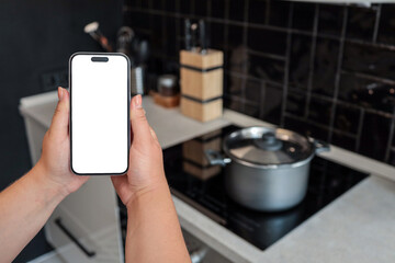 Two hands holding smartphone with blank screen in modern kitchen. Cooking pot on induction stove in...