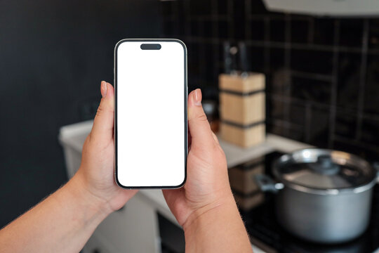 Two hands holding smartphone with blank screen in modern kitchen. Cooking pot on induction stove in the background, with black tiled backsplash and knife block visible