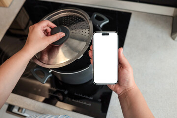Two hands holding smartphone with blank screen in modern kitchen. Cooking pot on induction stove in...