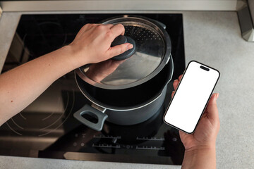 Person holding smartphone with blank screen in one hand while lifting pot lid with the other over...