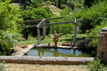 Garden pond with terracotta statue on stone platform, wooden pergola frame, and lush trees in a...