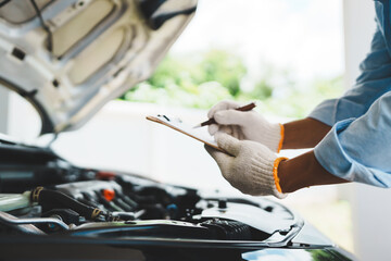 A mechanic wearing gloves inspects a car engine and keeps a checklist, focusing on maintenance and repair for the safety, reliability and optimum performance of the car in the auto repair shop.