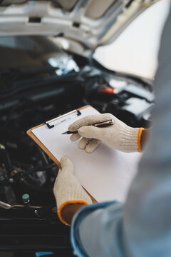 A mechanic wearing gloves inspects a car engine and keeps a checklist, focusing on maintenance and repair for the safety, reliability and optimum performance of the car in the auto repair shop.