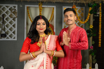 Portrait of a man in kurta pyjama and woman in saree, with gold jewellery, standing together with hands in namaste gesture.