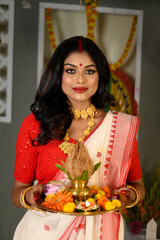 Portrait of young woman, adorned in traditional saree, complemented by gold jewellery, holding a plate filled with religious offerings.