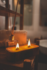 A handmade wooden candle with twin flames burning brightly on a bamboo shelf in a spa-style bathroom, accompanied by a rolled towel and glass diffuser, creating a serene and cozy atmosphere
