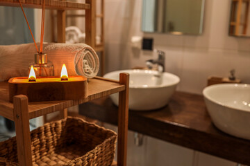 A handmade wooden candle with twin flames burning brightly on a bamboo shelf in a spa-style bathroom, accompanied by a rolled towel and glass diffuser, creating a serene and cozy atmosphere