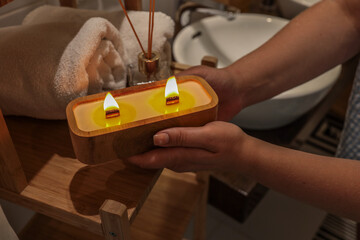 Woman holding a lit wooden wick candle in cozy bathroom with rolled towels and aroma diffuser. Warm...