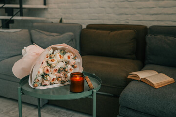 Bouquet of pale pink roses on a green table next to a lit candle in a cozy living room with soft...