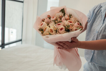 Close-up of a woman in a blue gingham outfit holding a bouquet of pastel roses wrapped in pink paper, set against a soft, minimal bedroom background with natural light