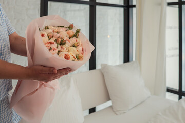 Close-up of a woman in a blue gingham outfit holding a bouquet of pastel roses wrapped in pink paper, set against a soft, minimal bedroom background with natural light