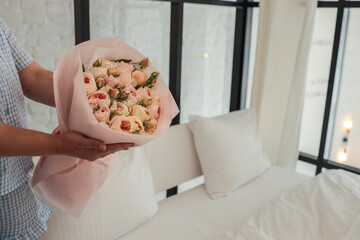 Close-up of a woman in a blue gingham outfit holding a bouquet of pastel roses wrapped in pink paper, set against a soft, minimal bedroom background with natural light
