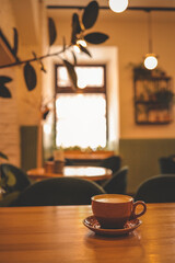 Warm and cozy coffee shop interior with a ceramic cup of cappuccino on a wooden table, soft lighting, blurred background with plants, and a relaxed, inviting atmosphere