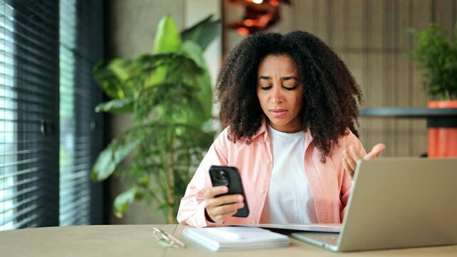 Beautiful African American freelancer, wearing a pink shirt, expresses intense frustration and confusion while using her smartphone at a contemporary office desk. - Powered by Adobe